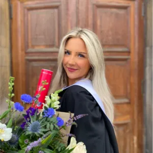 Photo of Rachael, smiling at the camera, wearing a black gown and holding a bunch of flowers.