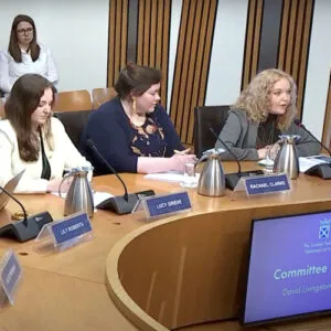 Dr Rebecca Mason, research & policy lead speaking at a panel with a group of women with name plates and microphones in front of them.