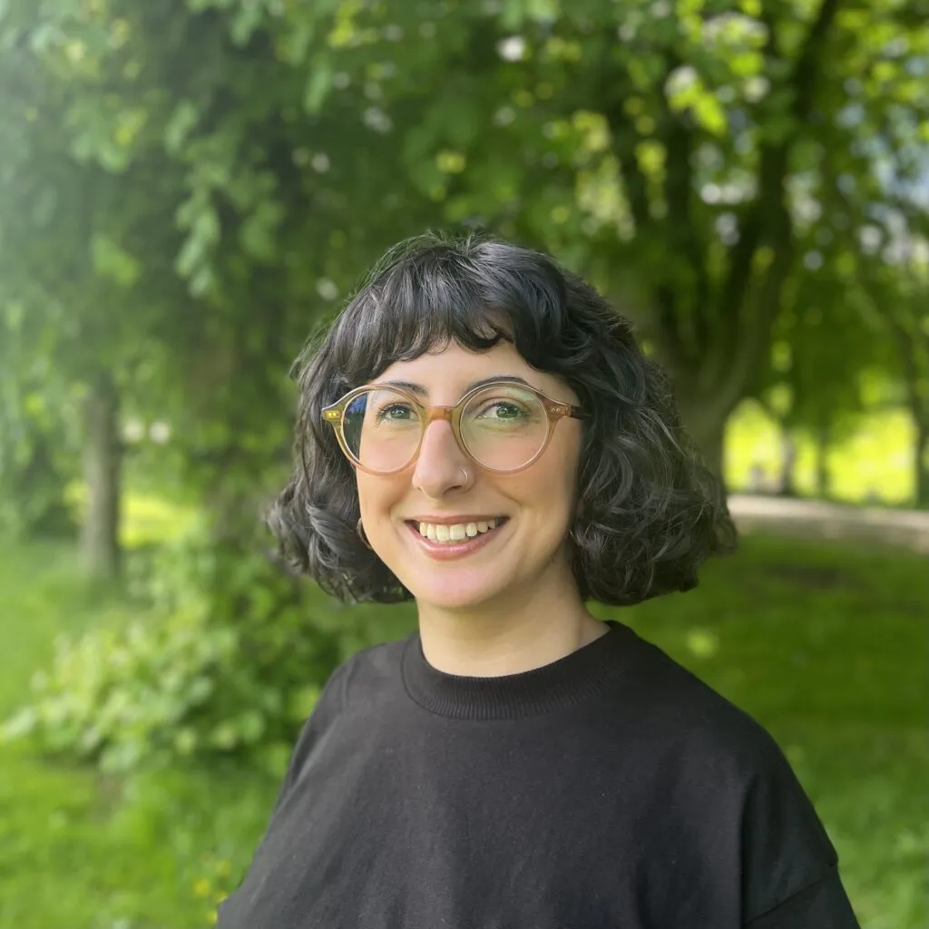 A photo of Samar, who has dark curly hair and glasses and is smiling at the camera. There are trees and grass in the background of the photo.