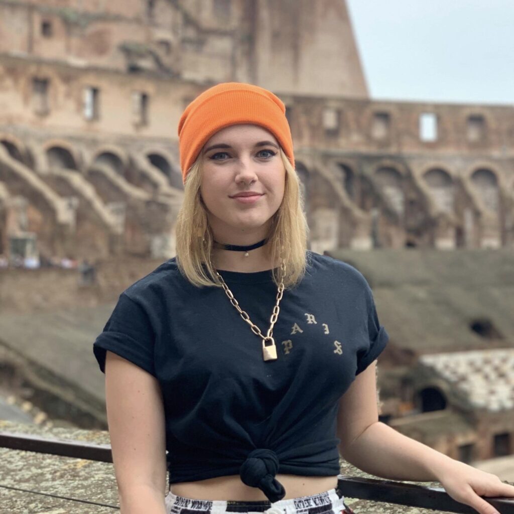 Cailyn McMahon looking at the camera wearing an orange hat, with a historic ruin behind her