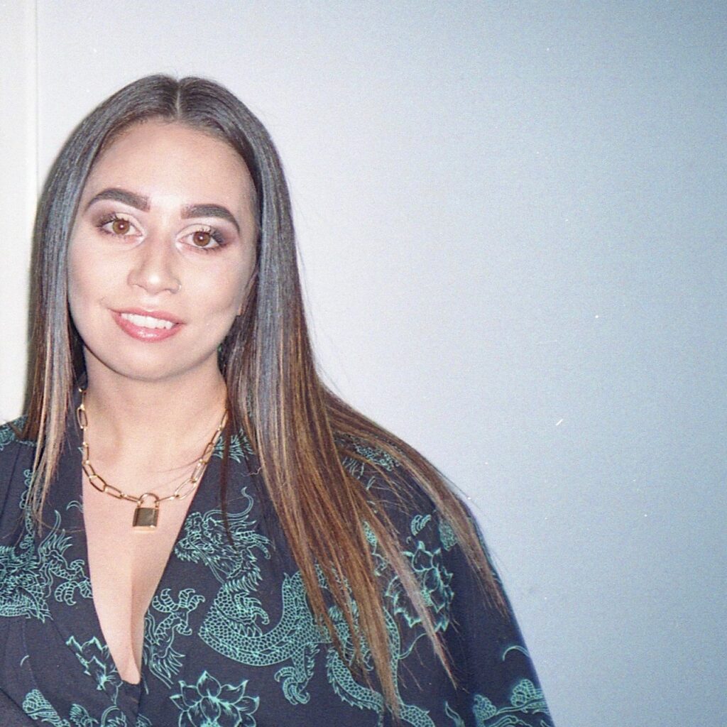 Denisha Killoh smiling at the camera against a plain background, with a bright flash from the camera on her face