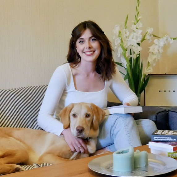 Emily Horgan smiling at the camera while sitting on a sofa holding a book and with a labrador beside her