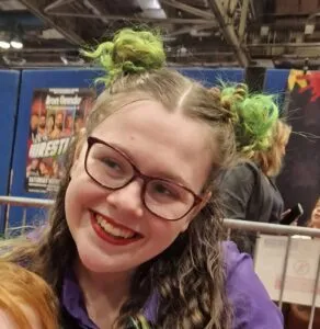 Kaydi has pale skin and her hair is dyed green and tied into space buns. She is wearing glasses and smiling.