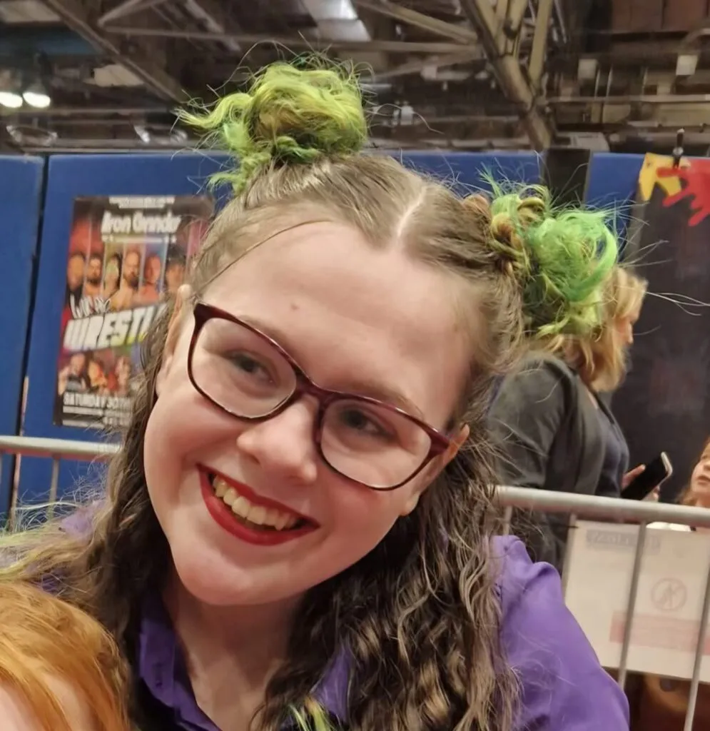 Kaydi has pale skin and her hair is dyed green and tied into space buns. She is wearing glasses and smiling.
