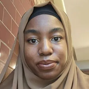 A young black woman, wearing a beige hijab, looking at the camera.