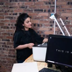 Sarah Naseer looking at something out of shot, with a brick wall behind her and a computer in the foreground