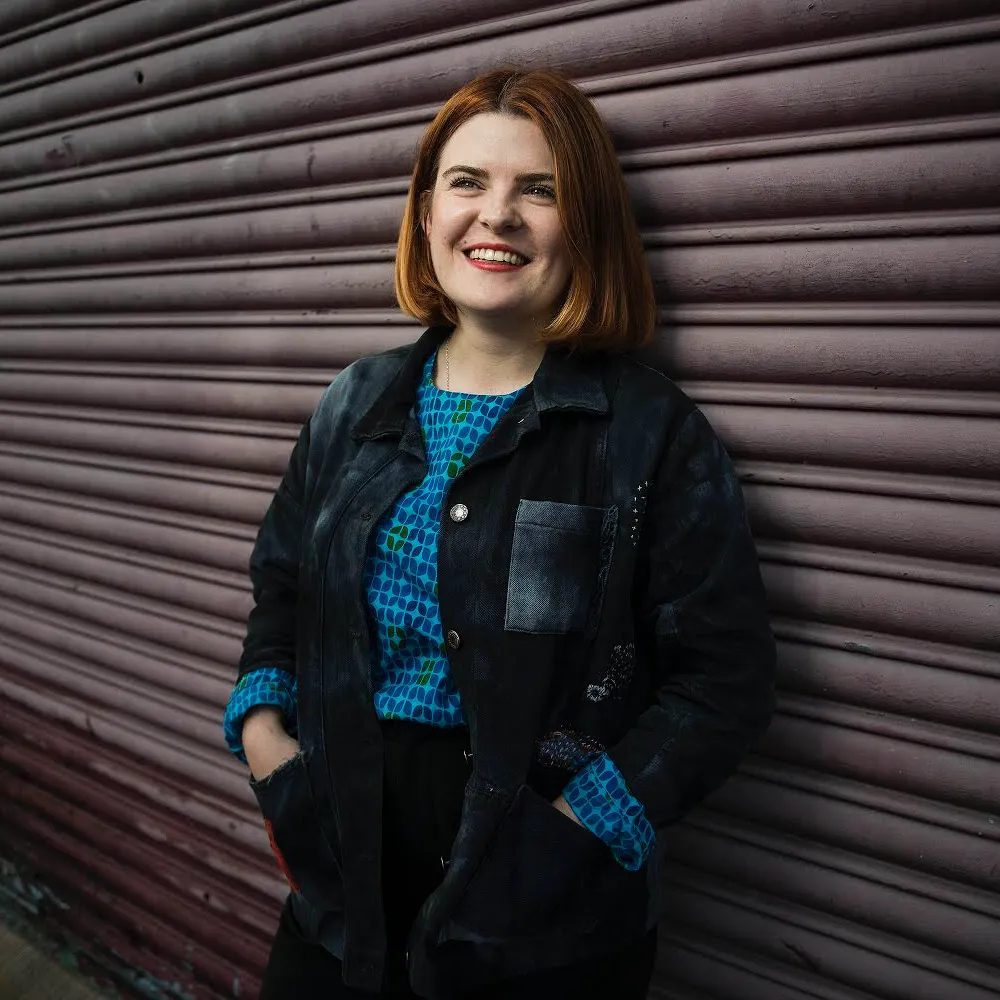 Siobhan McKenna smiling and looking off camera, leaning against an outside metal wall