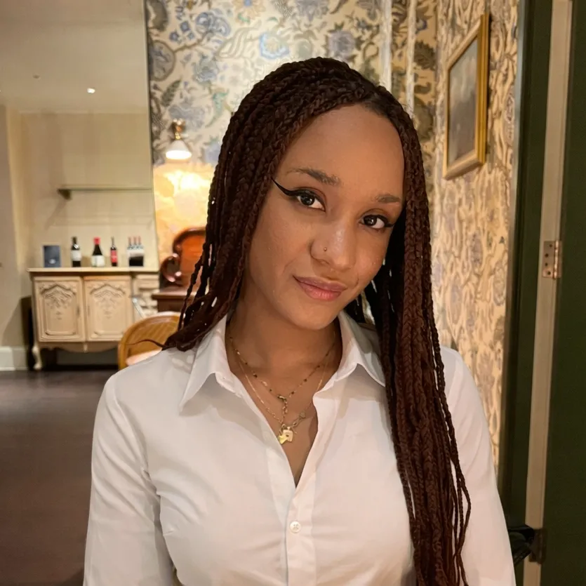 Stella has dark skin and long hair in braids. She is wearing a white shirt and sitting in a restaurant.