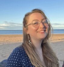 Gabrielle wears glasses and is smiling at the camera in front of a beach. She wears a blue top with white polkadots and dangly earrings.