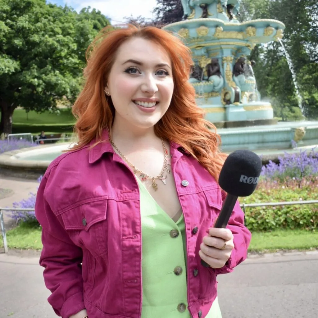 Alice Cruickshank smiling at the camera whilst holding a microphone, standing in front of a fountain.