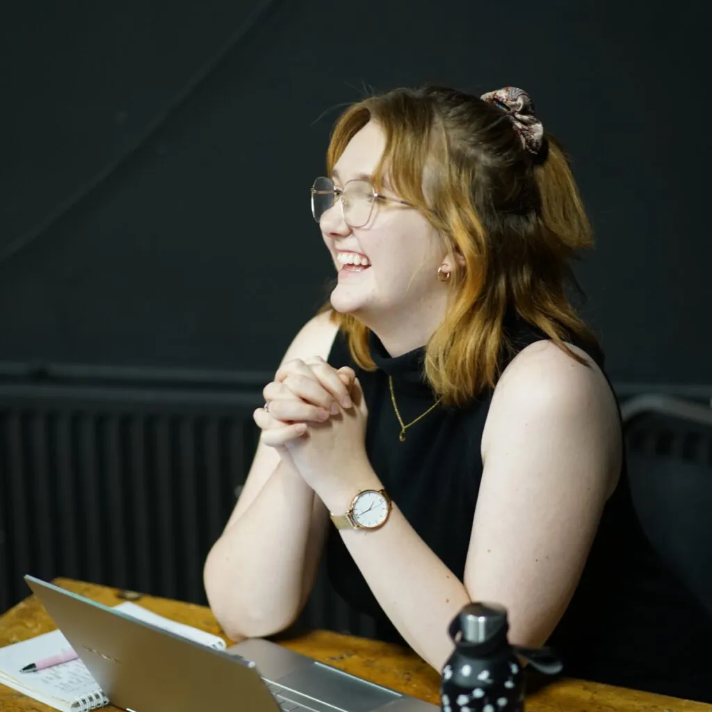 Emma smiling at someone off camera whilst leaning on a table.