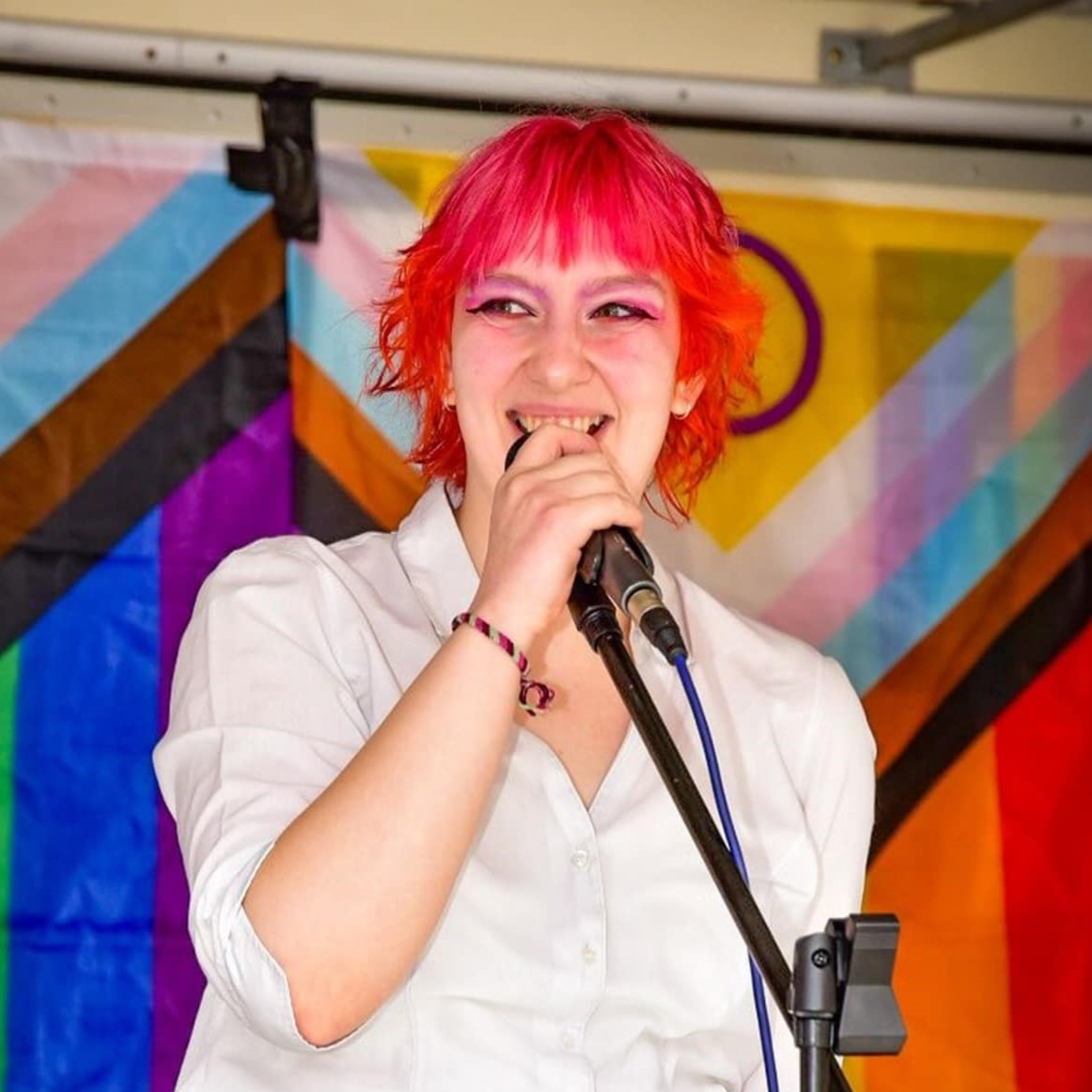 Emily smiling while holding a microphone in a mic stand, with pride flags in the background