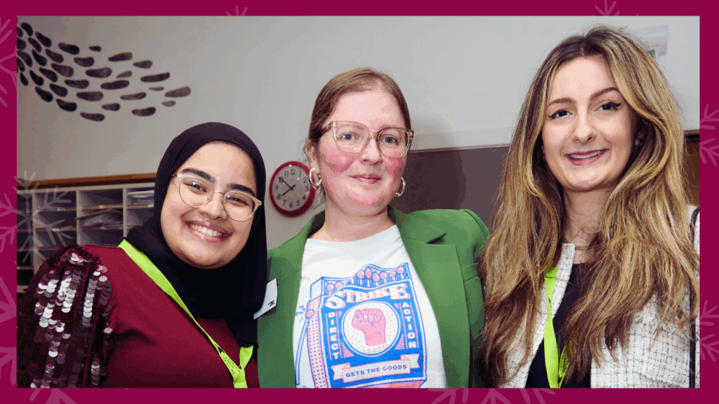 Three young women stand smiling together. One is wearing a hijab and glasses, another is wearing a green blazer with her hair up, and the third has her hair down.