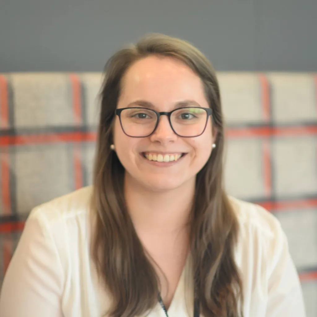 Photo of a young woman smiling at the camera, she has long brown hair and glasses. There is a beige, red and blue checkered pattern in the background.