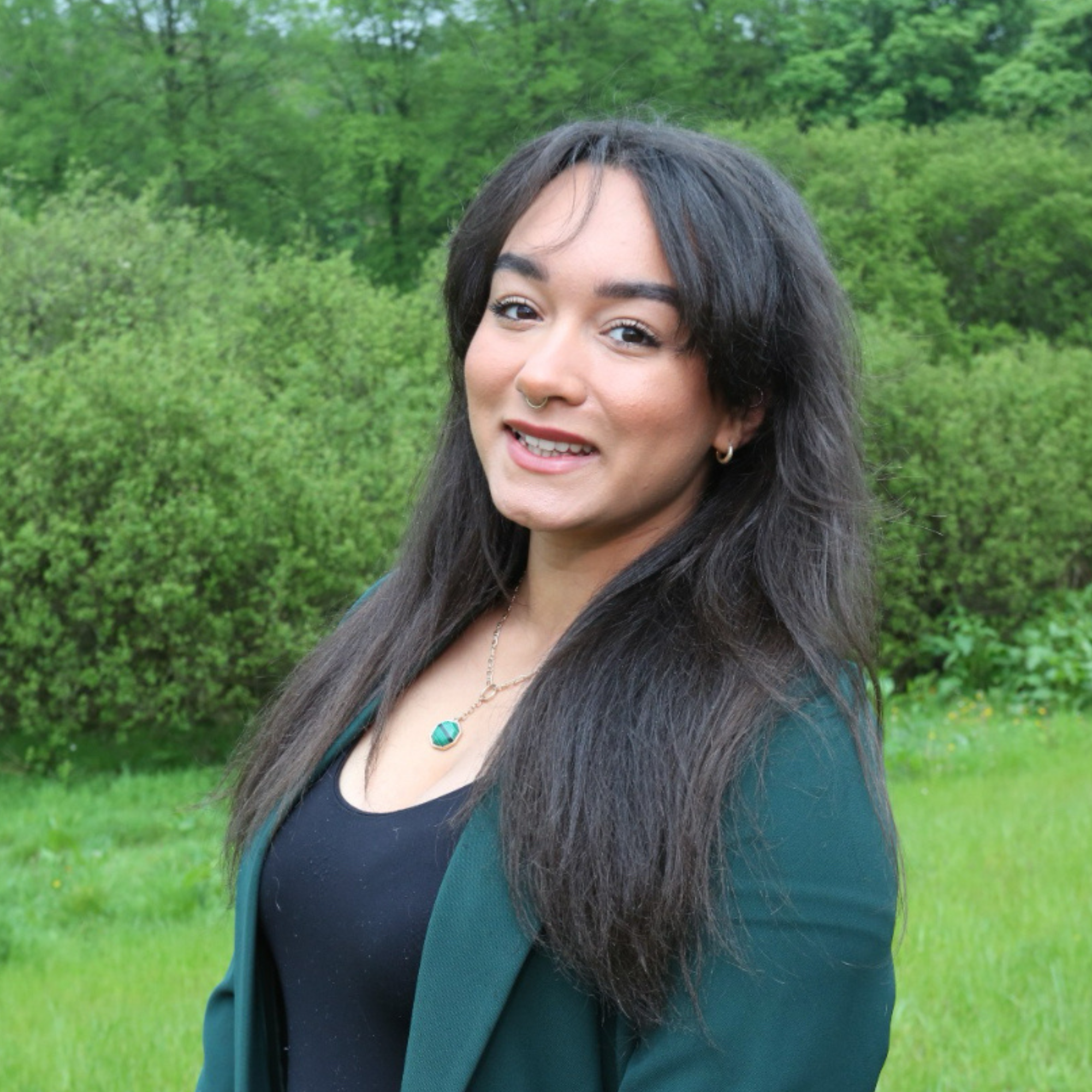 Maya has long black hair, and is smiling at the camera. There are green trees and grass behind here.