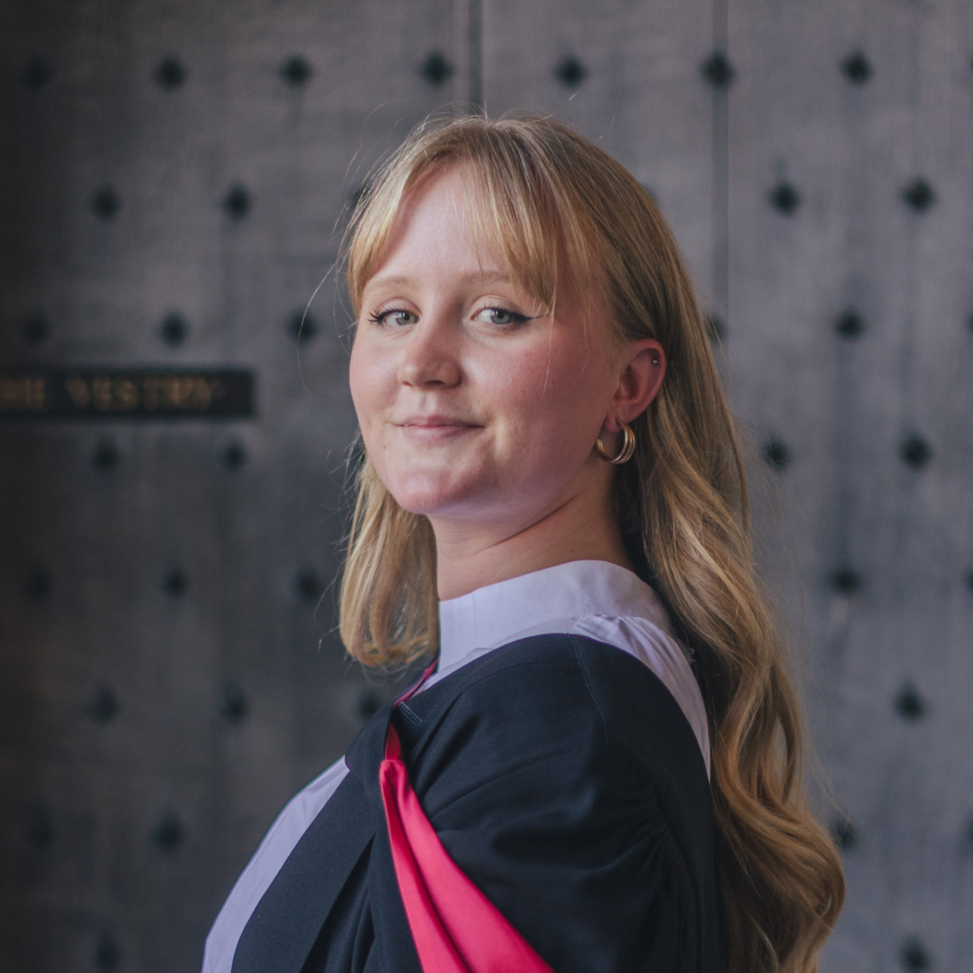 Yasmine has long blonde hair and is smiling at the camera, she is wearing graduation robes in black and red.