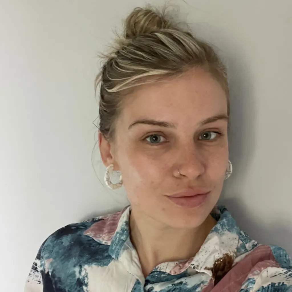 Photo of Natalie, who was blonde hair in a bun. She has hoop earrings, and is wearing a blue, pink, and white shirt.