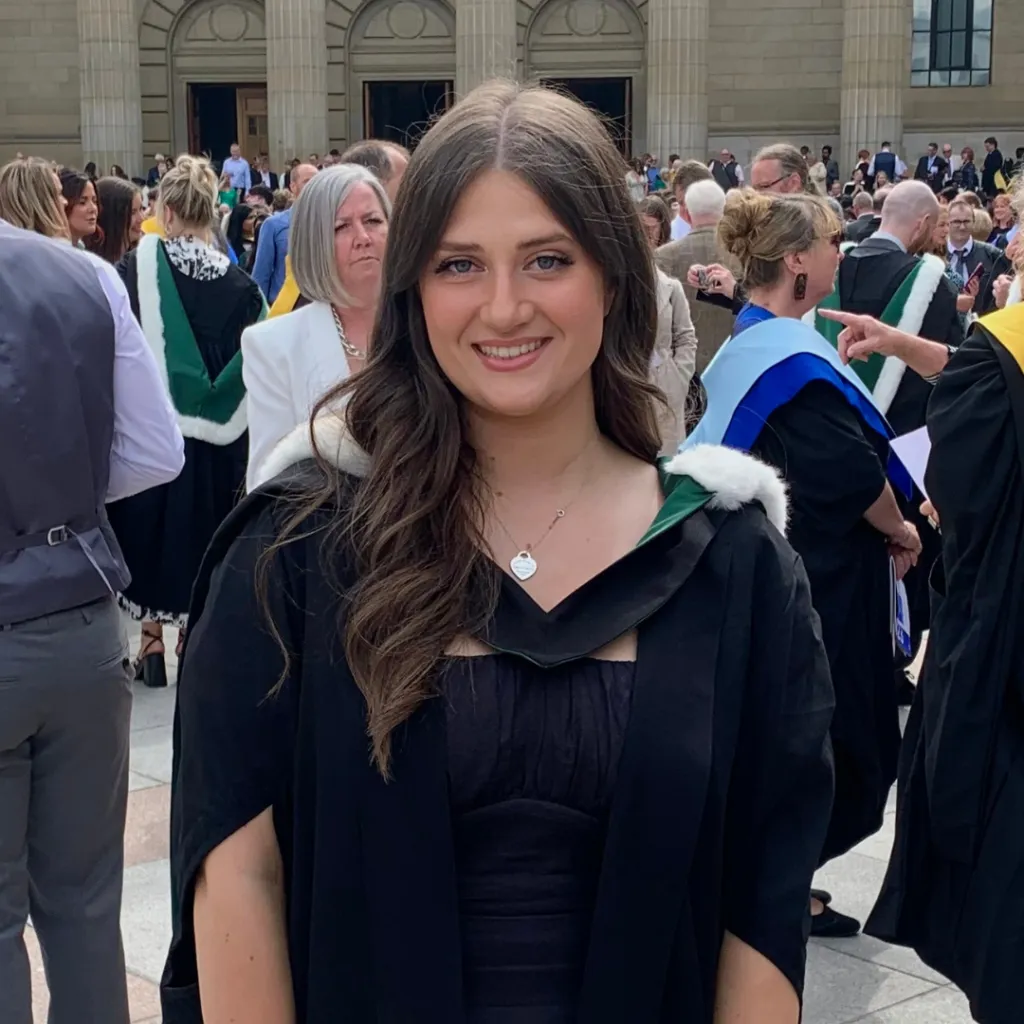 Photo of Lauren, who is a Community and Family worker. She has long brown hair and is smiling at the camera. Lauren is wearing a graduation gown, and there is a crowd of people behind her.