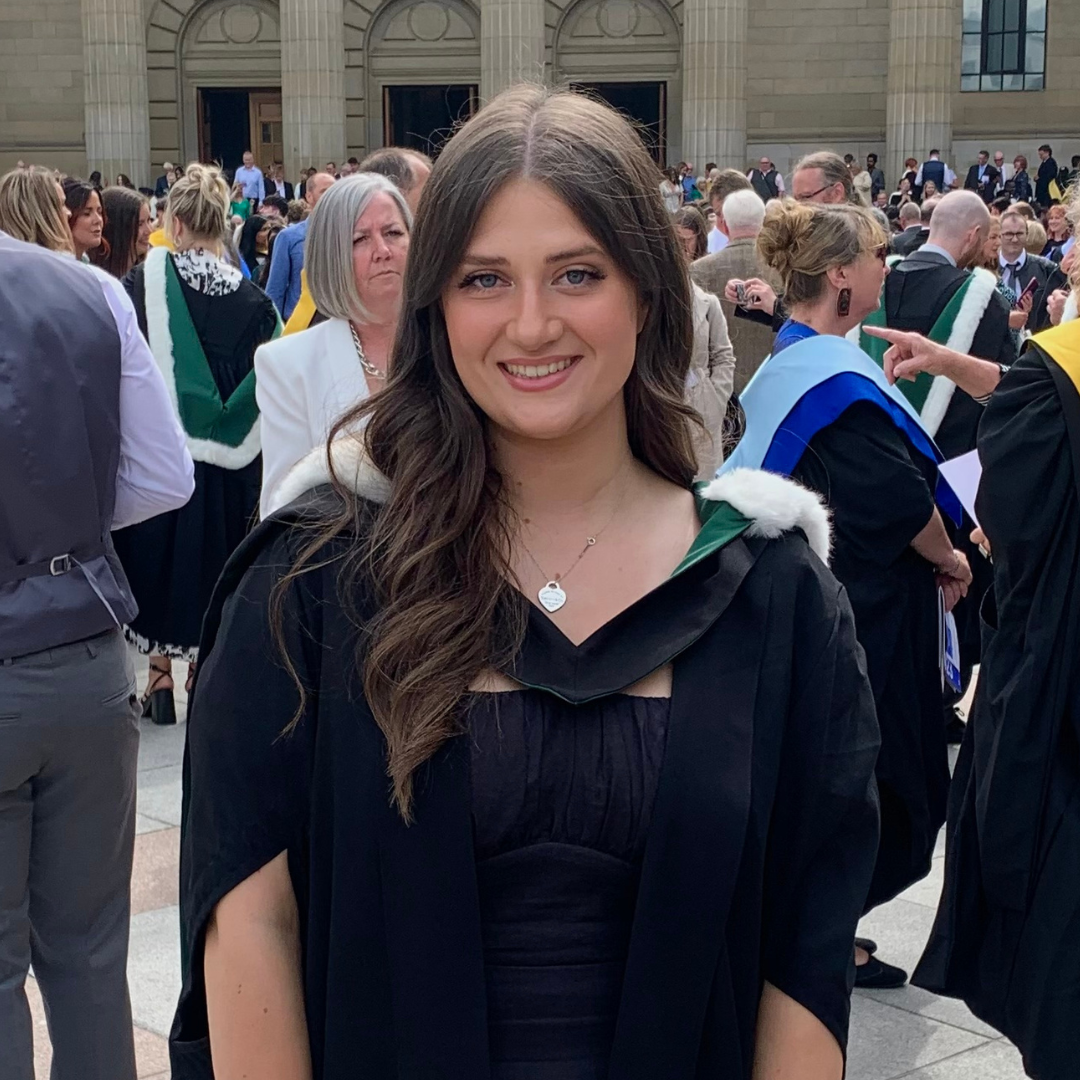 Photo of Lauren, who is a Community and Family worker. She has long brown hair and is smiling at the camera. Lauren is wearing a graduation gown, and there is a crowd of people behind her.