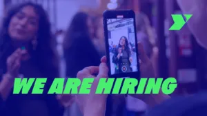 Graphic with text: We are hiring! Photo of a person interviewing and filming another person, via a mobile phone. There are arrow shapes and a Y logo in the corner.