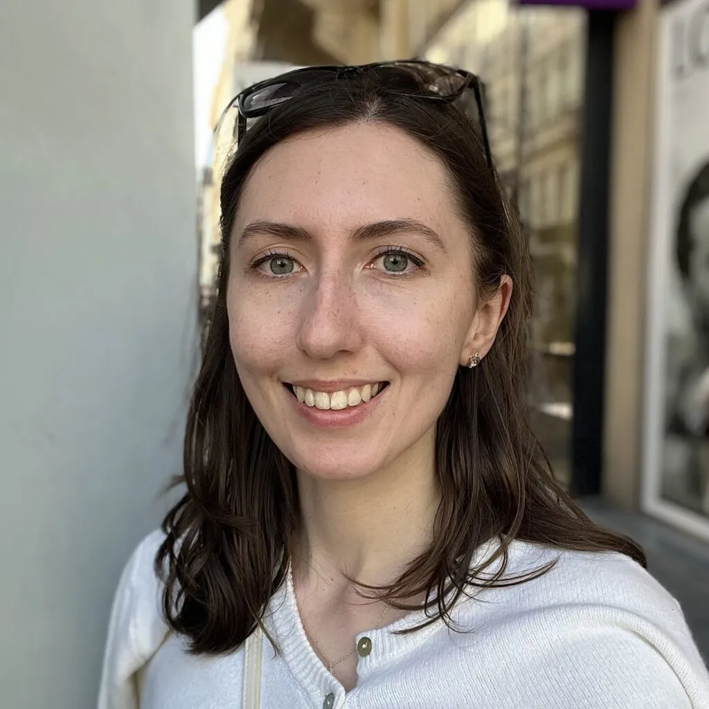 Alexandra has pale skin and dark hair. She is wearing a white shirt and has sunglasses on her head.