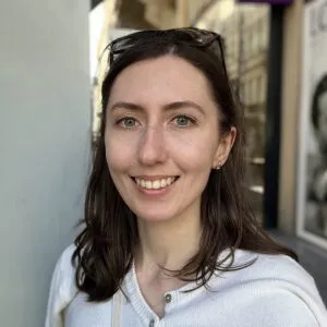 Alexandra has pale skin and dark hair. She is wearing a white shirt and has sunglasses on her head.