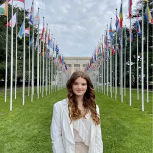 Beau is wearing a white outfit. She has pale skin and long brown hair. She is standing in front of a stately building surrounded by different flags.