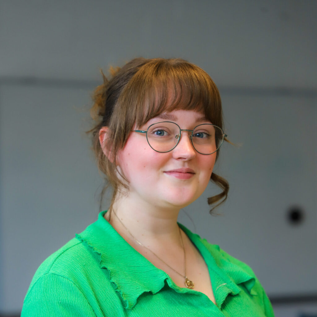 Bethany has pale skin and light brown hair with a fringe. She is wearing glasses and a bright gren blouse.