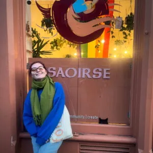 Chloe is wearing a blue jumper, a green scarf and glasses. They have pale skin and dark hair.