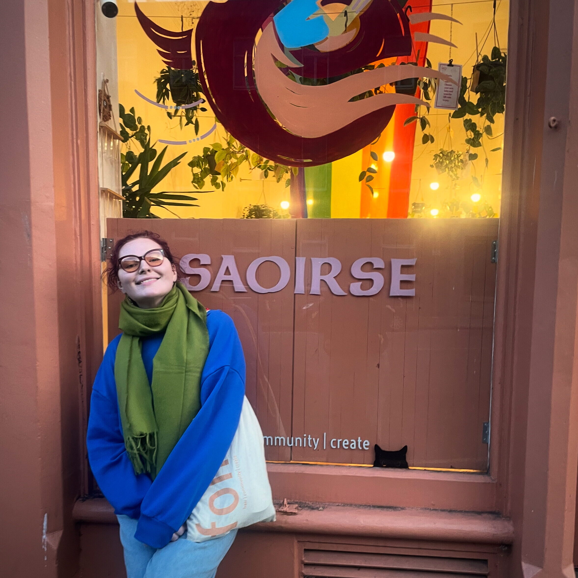 Chloe is wearing a blue jumper, a green scarf and glasses. They have pale skin and dark hair.