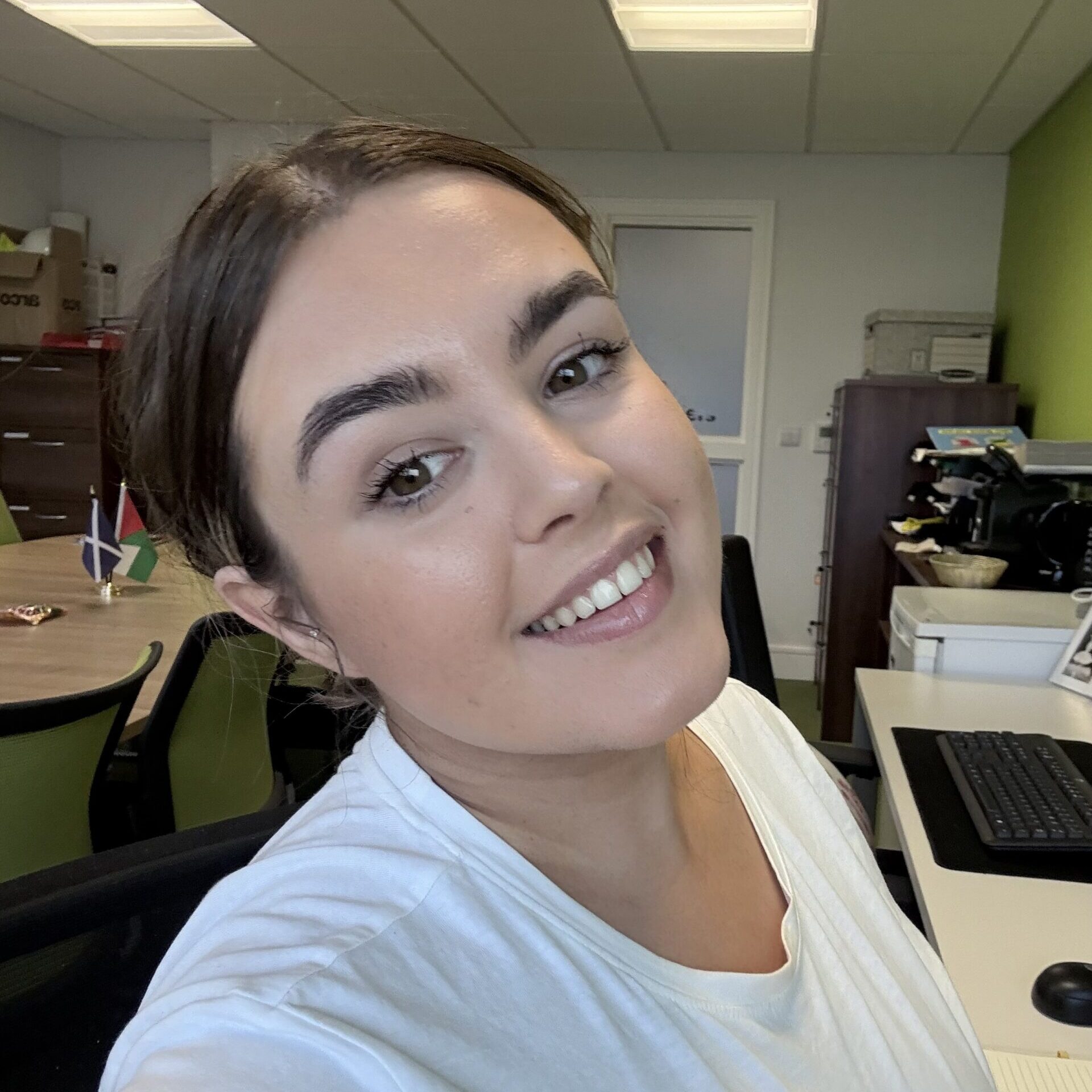 Chloe has pale skin and dark hair tied back. She is wearing a white t-shirt and appears to be in an office.