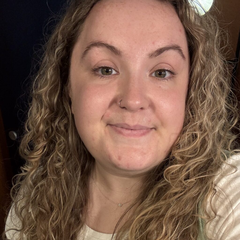 Emily has pale skin and curly blonde hair. She is smiling at the camera.