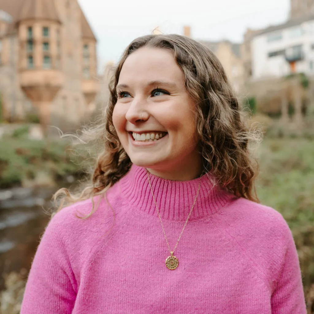 Emma has pale skin and curly blonde hair. She is in Dean billage in Edinburgh and is wearing a pink jumper.