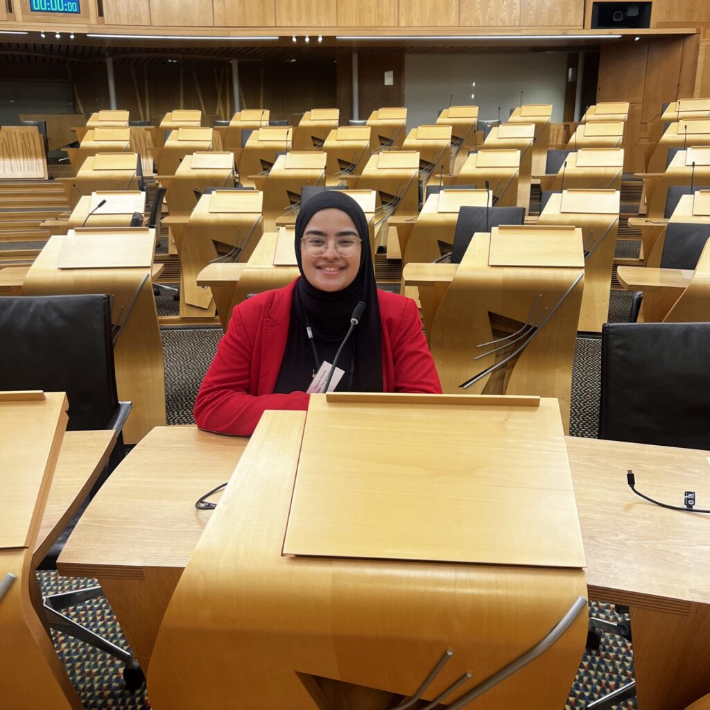 Fatemah is wearing a black hijab and a red blazer. She is sitting in the Scottish Parliament debating chamber.