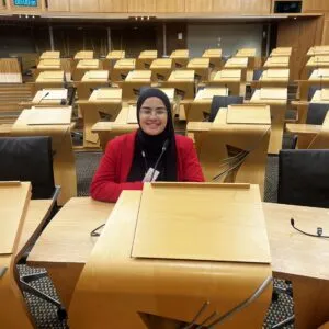 Fatemah is wearing a black hijab and a red blazer. She is sitting in the Scottish Parliament debating chamber.