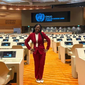 Favour has dark skin and long dark hair. She is wearing a matching red suit and is standing in a government building.