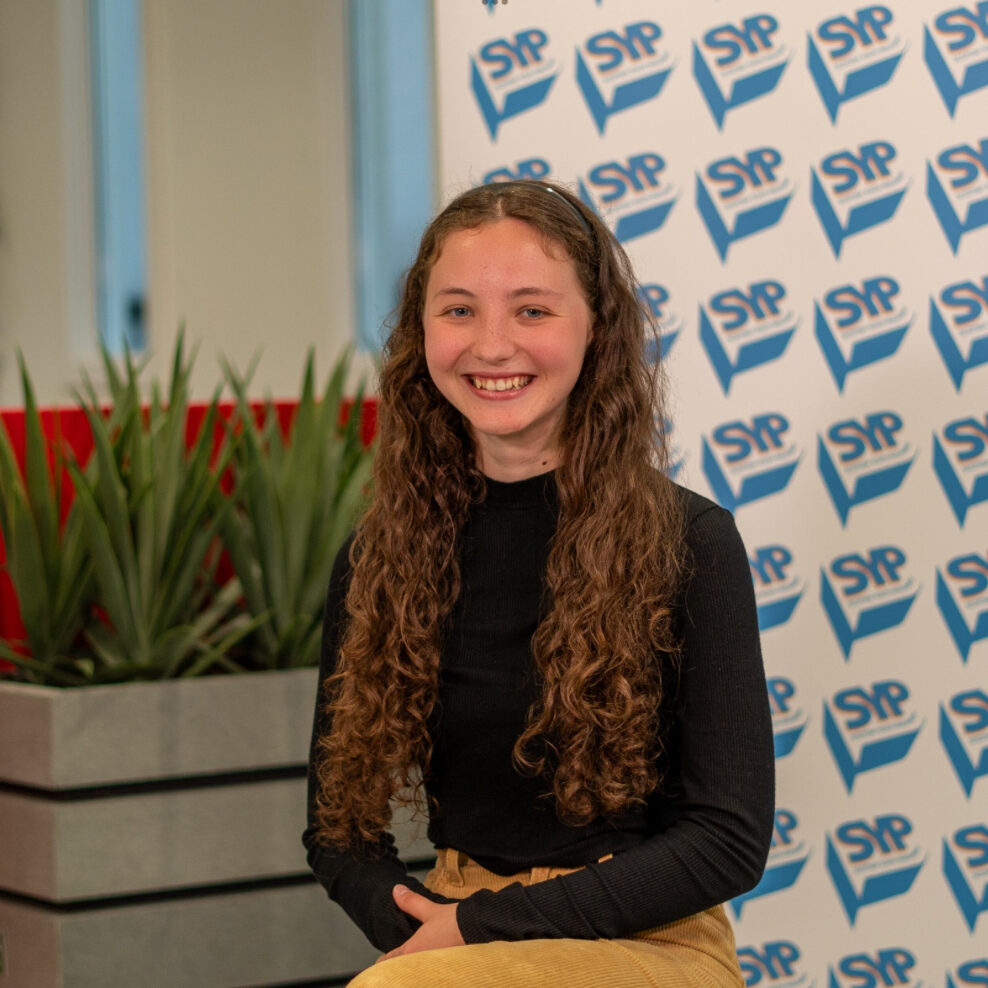 Islay has pale skin and long, curly brown hair. She is sitting in front of a Scottish Youth Parliament sign.