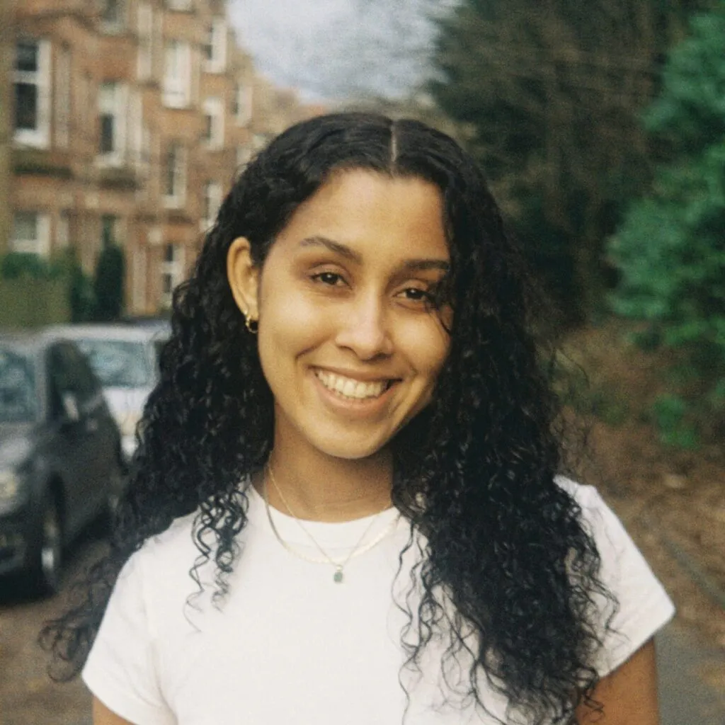 Jodie has brown skin and long curly black hair. She is wearing a white t-shirt and smiling at the camera.