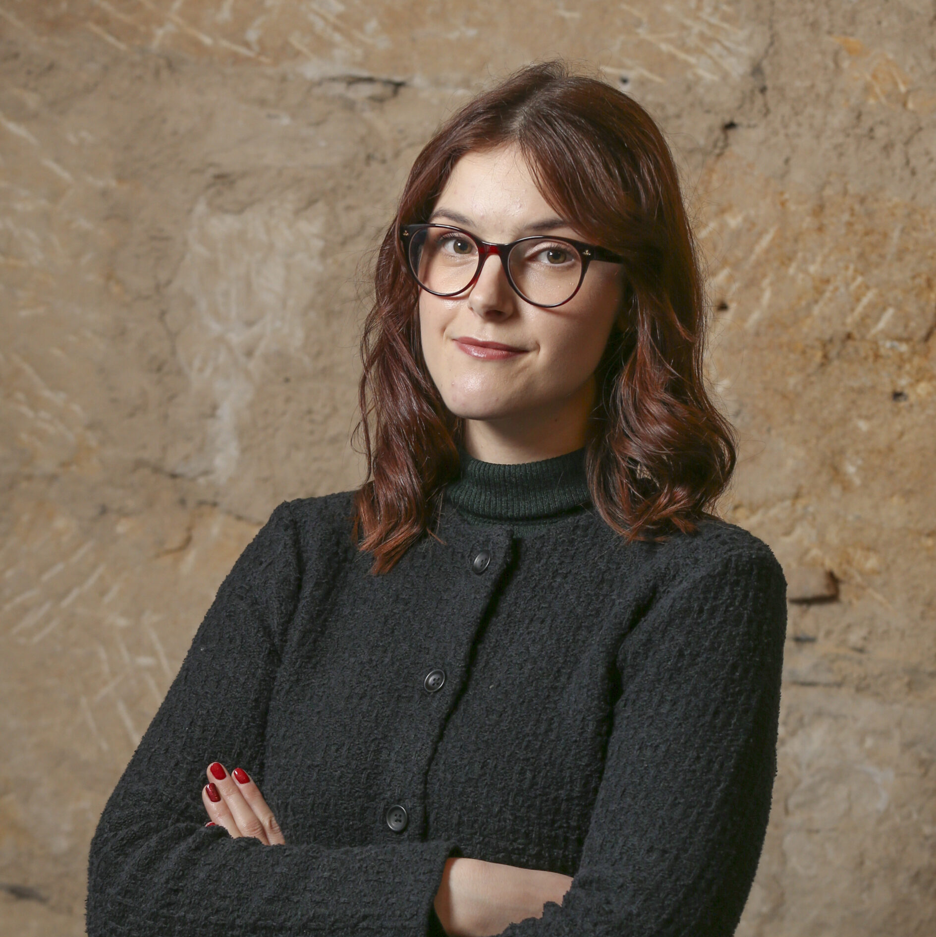 Laura has pale skin and red hair. She is wearing glasses and a black jumper.