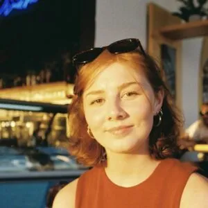 Leah has short red hair and pale skin. She is wearing a red top and sunglasses on her head.