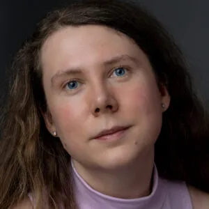 Lucy Beth has pale skin, dark curly hair and blue eyes. She is wearing a lilac top.