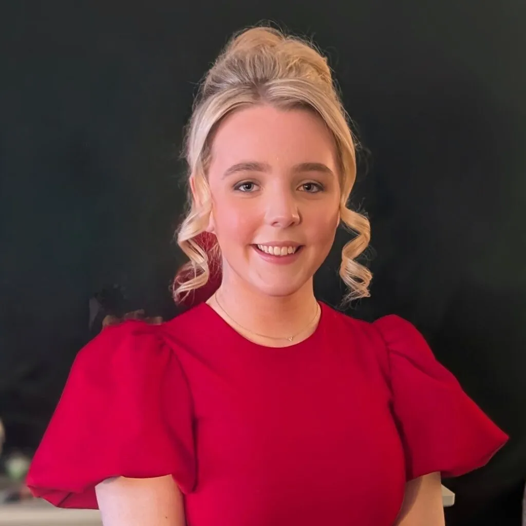 Orlagh has pale skin and blonde hair tied into a bun. She is wearing a red blouse.