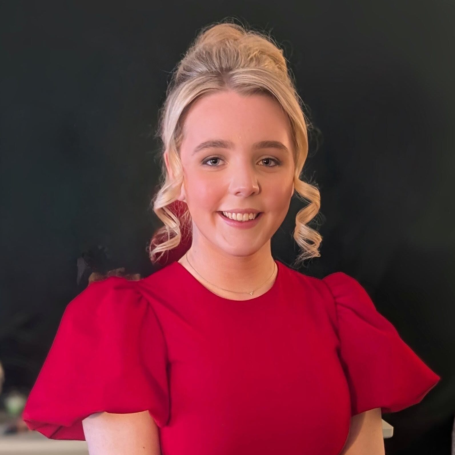 Orlagh has pale skin and blonde hair tied into a bun. She is wearing a red blouse.