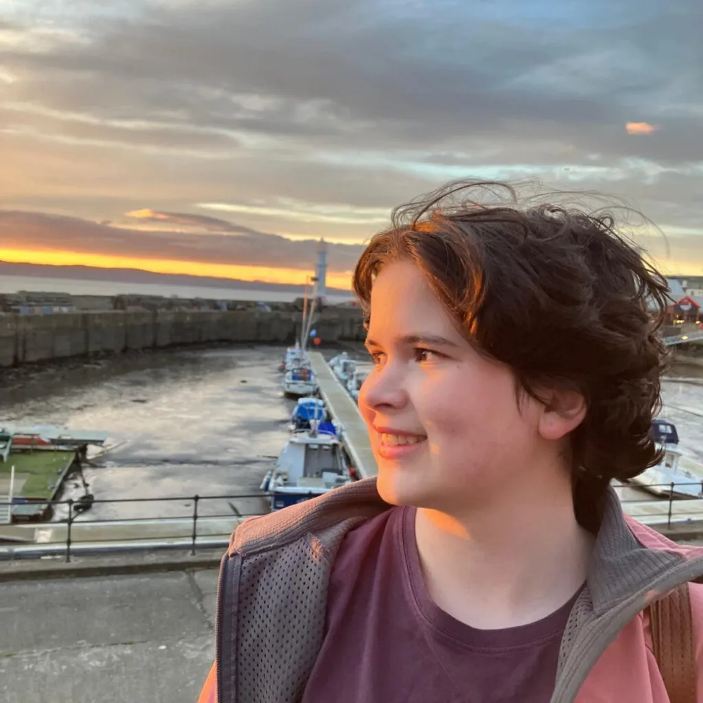 Rosa has pale skin and short dark hair. She is standing in front of a harbour at sunset.
