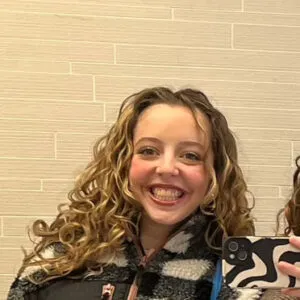 Mirin has pale skin and long blonde curly hair. She is wearing a black and white checkered fleece