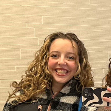 Mirin has pale skin and long blonde curly hair. She is wearing a black and white checkered fleece