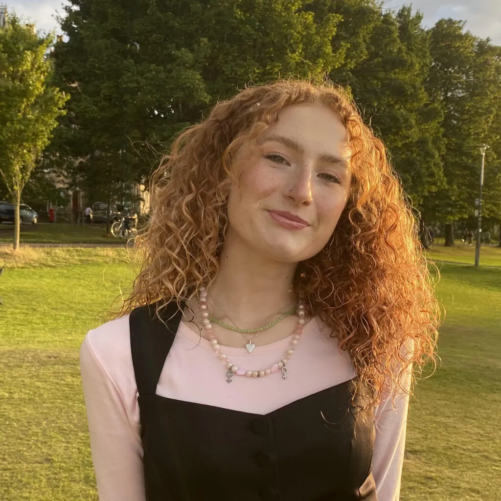 Matilda has pale skin and curly red hair. She is sitting in a park.
