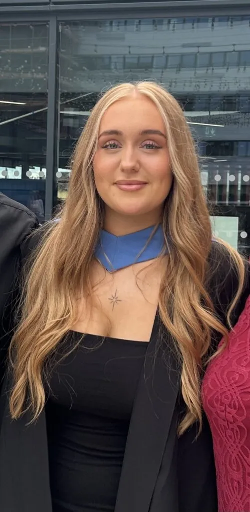 Natalie has tanned skin and long blonde hair. She is wearing a graduation gown.