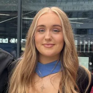 Natalie has tanned skin and long blonde hair. She is wearing a graduation gown.