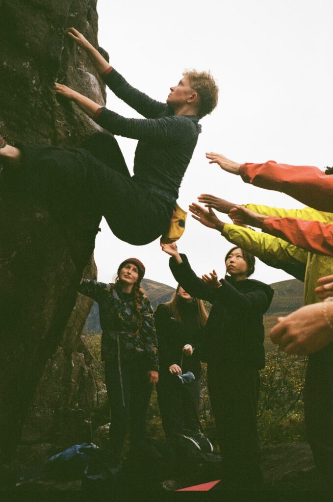A person with short hair is climbing a rockface, and hands reach out to support them